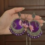 "Hand holding a pair of intricately designed silver and purple earrings with floral resin details, accompanied by a matching pendant necklace."