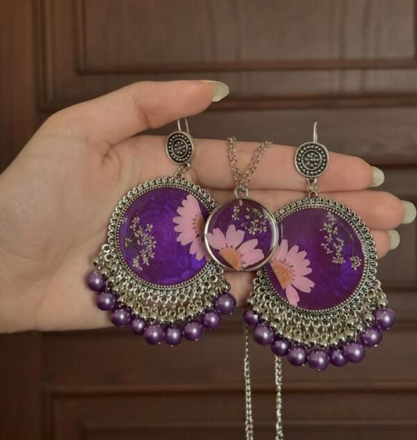 "Hand holding a pair of intricately designed silver and purple earrings with floral resin details, accompanied by a matching pendant necklace."
