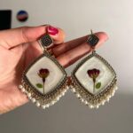 "Hand holding a pair of handcrafted vintage-style earrings with embedded real rosebuds, pearl embellishments, and intricate metallic detailing."