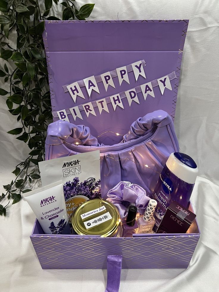 A beautifully arranged purple-themed birthday gift box containing skincare products, a satin bag, a scrunchie, a perfume, nail polish, and a "Memories Jar," with a "Happy Birthday" banner attached to the lid.