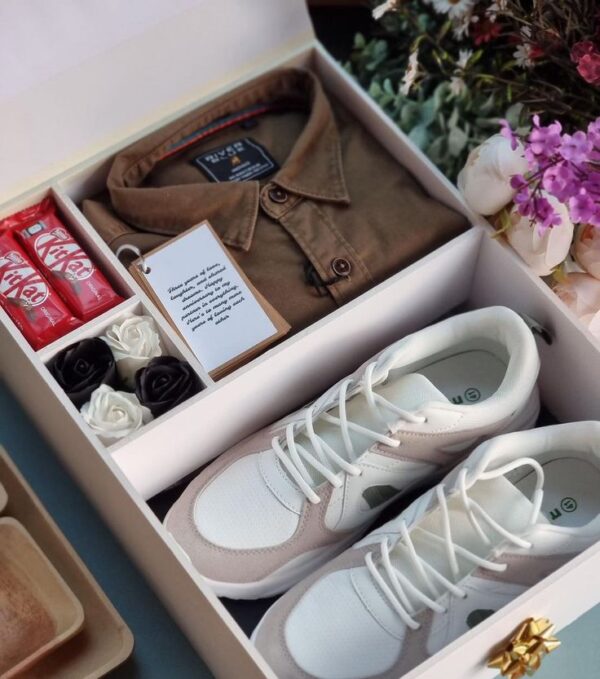A neatly arranged gift box containing a folded brown shirt, a pair of white and beige sneakers, two KitKat chocolate bars, artificial black and white roses, and a small note card with a heartfelt message.