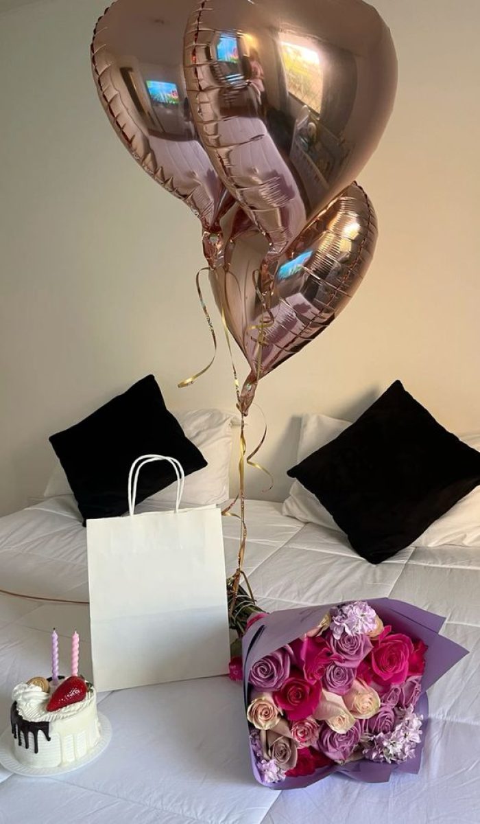A beautifully decorated bed with a white bedsheet, featuring heart-shaped rose gold balloons, a white gift bag, a bouquet of pink and purple roses, and a small cake with two candles.